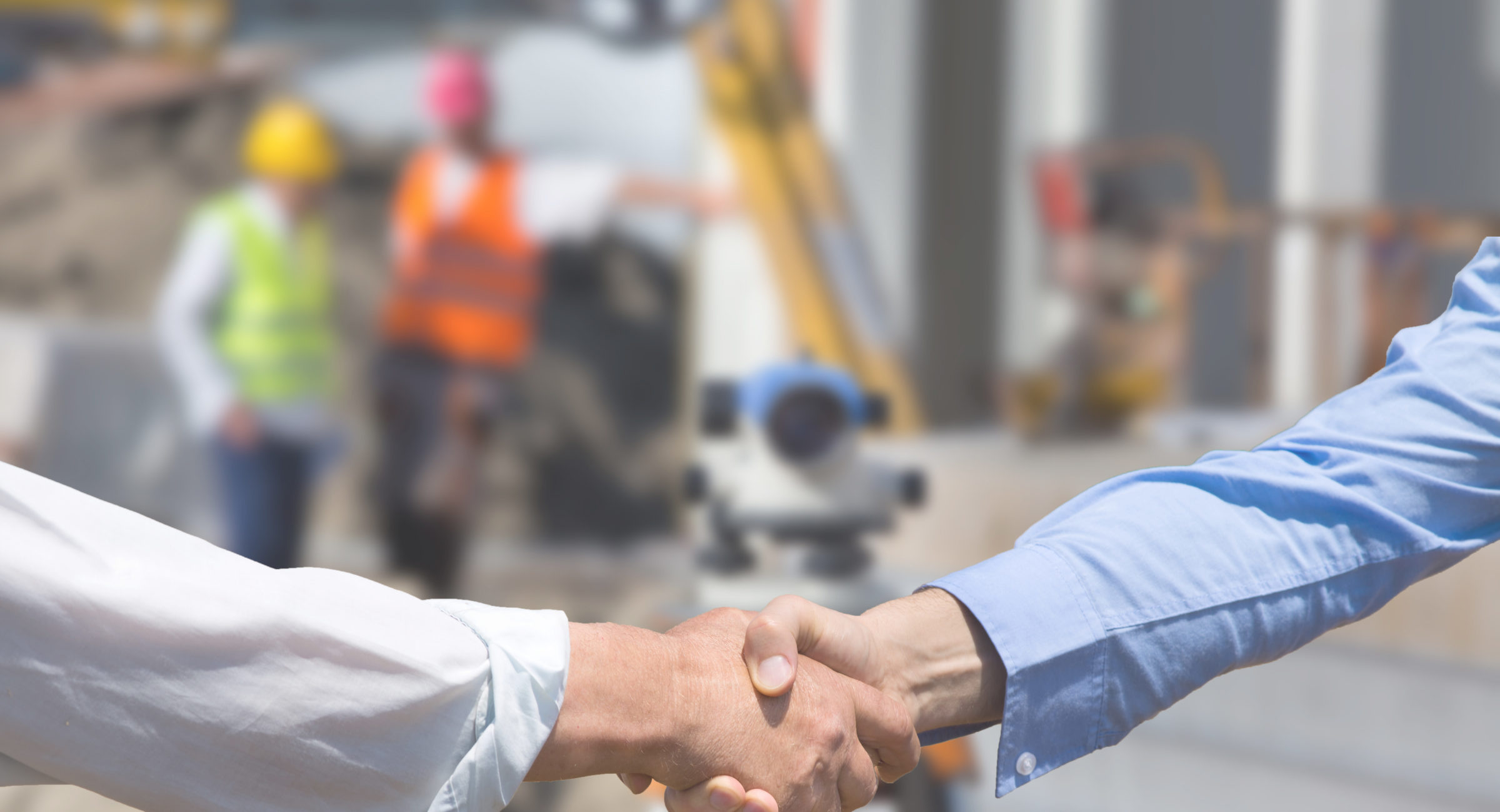 Two business people shaking hands in front of leveling instrument on construction site with workers in background